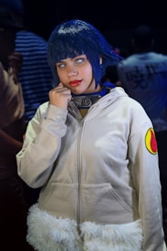 A person in a costume featuring a blue wig and a beige jacket with a symbol resembling a swirl. The costume includes a necklace resembling a headband with a metal plate bearing a symbol. The person is posing with a hand near the chin and has a focused expression.