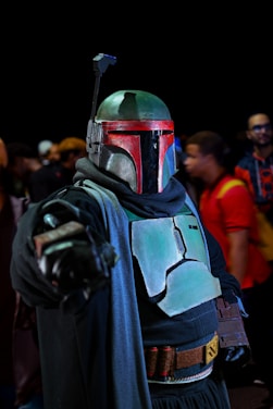 A person dressed in a sci-fi bounty hunter costume, featuring a helmet with a horizontal black visor and armor plates in a dark green hue. They are holding a replica blaster and have a cloak draped around their shoulders. The background includes several out-of-focus individuals, creating a sense of a crowded event or convention.