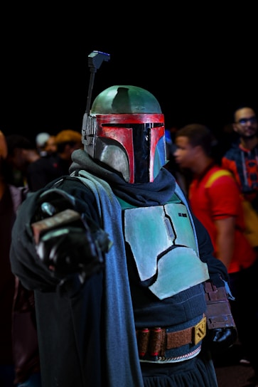 A person dressed in a sci-fi bounty hunter costume, featuring a helmet with a horizontal black visor and armor plates in a dark green hue. They are holding a replica blaster and have a cloak draped around their shoulders. The background includes several out-of-focus individuals, creating a sense of a crowded event or convention.