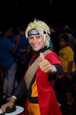 A person is dressed in a detailed cosplay outfit with a spiky blonde wig and a headband featuring a symbol. They are wearing a red and black costume with orange accents and giving a thumbs-up gesture, smiling. The background includes people in casual clothing, indicating a public event or convention.