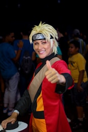 A person is dressed in a detailed cosplay outfit with a spiky blonde wig and a headband featuring a symbol. They are wearing a red and black costume with orange accents and giving a thumbs-up gesture, smiling. The background includes people in casual clothing, indicating a public event or convention.