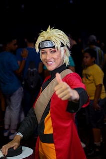 A person is dressed in a detailed cosplay outfit with a spiky blonde wig and a headband featuring a symbol. They are wearing a red and black costume with orange accents and giving a thumbs-up gesture, smiling. The background includes people in casual clothing, indicating a public event or convention.