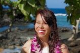 A woman smiles brightly while wearing a floral lei around her neck. She is outdoors with tropical trees and the ocean in the background, suggesting a sunny beach setting.