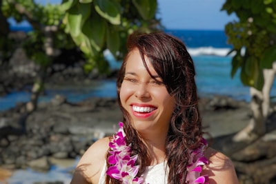 A woman smiles brightly while wearing a floral lei around her neck. She is outdoors with tropical trees and the ocean in the background, suggesting a sunny beach setting.