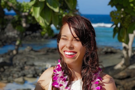 A woman smiles brightly while wearing a floral lei around her neck. She is outdoors with tropical trees and the ocean in the background, suggesting a sunny beach setting.