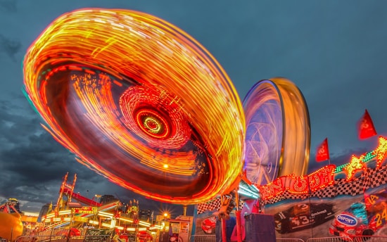 Bright, colorful lights spin rapidly, creating circular patterns in an amusement park setting. Various rides and attractions, including a Ferris wheel, are visible in the background against a twilight sky.
