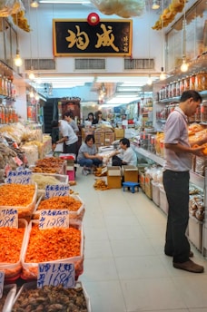 A bustling market stall filled with various dried goods in transparent containers and bags, likely seafood or herbs, with people actively engaged in packing and handling the goods. The area is well-lit with hanging light bulbs, and a large sign with Chinese characters is prominently displayed above. The floor is tiled, and there are boxes and stools scattered around.