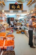 A bustling market stall filled with various dried goods in transparent containers and bags, likely seafood or herbs, with people actively engaged in packing and handling the goods. The area is well-lit with hanging light bulbs, and a large sign with Chinese characters is prominently displayed above. The floor is tiled, and there are boxes and stools scattered around.