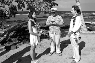 Three individuals are standing on a beach near the ocean. Two of them, a man and a woman, are facing each other and appear to be engaged in a ceremony, likely a wedding, as indicated by the leis they are wearing. The other man, who is wearing a patterned shirt and a white cap, appears to be officiating the ceremony. Palm trees and coastal rocks are visible in the background.