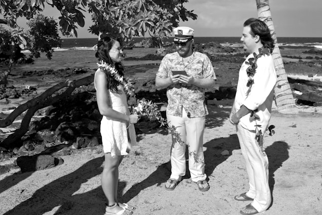 Three individuals are standing on a beach near the ocean. Two of them, a man and a woman, are facing each other and appear to be engaged in a ceremony, likely a wedding, as indicated by the leis they are wearing. The other man, who is wearing a patterned shirt and a white cap, appears to be officiating the ceremony. Palm trees and coastal rocks are visible in the background.