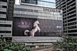 A large advertising billboard is mounted on the side of a gray, modern high-rise building. The ad features a white horse rearing on its hind legs against a dark, ornate background. Text on the billboard includes the words 'WINFIELD' and 'A WORLD APART', with additional text and contact details in smaller fonts. The building's facade has a grid-like pattern, and there are trees and more skyscrapers visible in the vicinity.