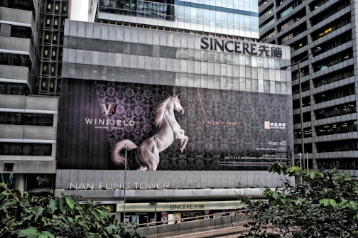 A large advertising billboard is mounted on the side of a gray, modern high-rise building. The ad features a white horse rearing on its hind legs against a dark, ornate background. Text on the billboard includes the words 'WINFIELD' and 'A WORLD APART', with additional text and contact details in smaller fonts. The building's facade has a grid-like pattern, and there are trees and more skyscrapers visible in the vicinity.