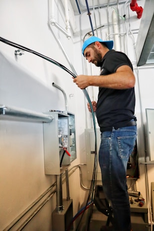 A man wearing a blue cap is working in a utility room, handling electrical wiring and a control panel. He stands on a step stool, focusing intently on the task. The background features various pipes, electrical conduits, and panels.