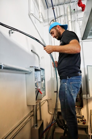 A man wearing a blue cap is working in a utility room, handling electrical wiring and a control panel. He stands on a step stool, focusing intently on the task. The background features various pipes, electrical conduits, and panels.
