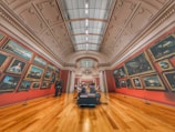 A grand art gallery with high ceilings and ornate decorations features numerous framed paintings on red walls. Two people are observing the artwork, and another person is sitting on a bench in the center, possibly reading about the exhibits. The warm wooden flooring contrasts with the cool tones of the paintings.