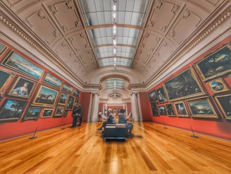 A grand art gallery with high ceilings and ornate decorations features numerous framed paintings on red walls. Two people are observing the artwork, and another person is sitting on a bench in the center, possibly reading about the exhibits. The warm wooden flooring contrasts with the cool tones of the paintings.