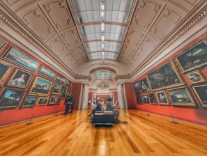 A grand art gallery with high ceilings and ornate decorations features numerous framed paintings on red walls. Two people are observing the artwork, and another person is sitting on a bench in the center, possibly reading about the exhibits. The warm wooden flooring contrasts with the cool tones of the paintings.