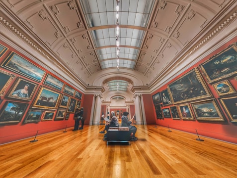 A grand art gallery with high ceilings and ornate decorations features numerous framed paintings on red walls. Two people are observing the artwork, and another person is sitting on a bench in the center, possibly reading about the exhibits. The warm wooden flooring contrasts with the cool tones of the paintings.