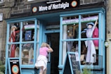 A quaint shopfront named 'Wave of Nostalgia' with large blue windows and a door. The window displays colorful historical illustrations of women, possibly related to women's suffrage, with banners stating 'Votes' and 'Women 100 years'. A person wearing a pink top and white skirt is peering into the store, which seems to have various nostalgic items on display, including books and dollhouses.
