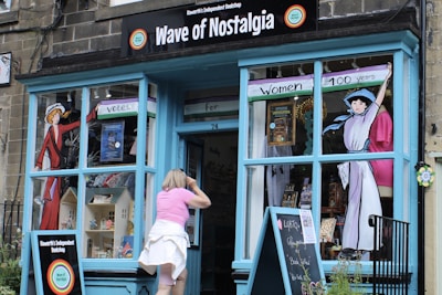 A quaint shopfront named 'Wave of Nostalgia' with large blue windows and a door. The window displays colorful historical illustrations of women, possibly related to women's suffrage, with banners stating 'Votes' and 'Women 100 years'. A person wearing a pink top and white skirt is peering into the store, which seems to have various nostalgic items on display, including books and dollhouses.