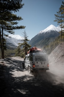 A rugged vehicle travels on a dusty mountain road with snow-capped peaks in the distance. The vehicle is loaded with gear secured on the roof, heading into a densely forested area under a clear blue sky.