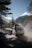 A rugged vehicle travels on a dusty mountain road with snow-capped peaks in the distance. The vehicle is loaded with gear secured on the roof, heading into a densely forested area under a clear blue sky.