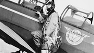 A man with a helmet and flight gear is standing next to an airplane, holding onto the side of the aircraft. The airplane has a logo that includes an image of a globe and wings with text written on it.