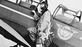 A man with a helmet and flight gear is standing next to an airplane, holding onto the side of the aircraft. The airplane has a logo that includes an image of a globe and wings with text written on it.