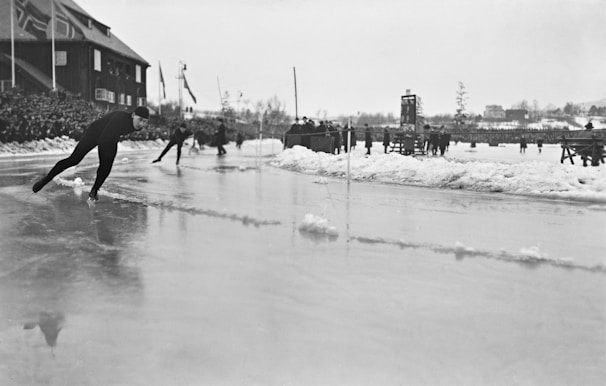 Several skaters are competing on an outdoor ice rink surrounded by snow. The skaters, dressed in dark winter attire, glide along the icy surface, while spectators gather around the perimeter. Flags are visible in the background, alongside a structure resembling a scoreboard. The scene is set under an overcast sky, contributing to the monochromatic atmosphere.
