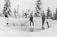 Two individuals are working in a snowy landscape, cutting and moving blocks of ice. They are dressed in winter clothing and are using tools such as a large saw and a shovel. The background features snow-covered trees, creating a serene winter environment.