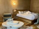 A modern bedroom with a messy bed on a wooden frame, a contemporary green armchair, a white abstract coffee table with a laptop on it, and a beige wall with a single illuminated wall sconce. A folded green blanket is also seen on the bed, and a patterned rug is underneath the table.