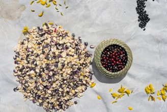 A collection of various seeds and grains spread on a light-colored surface, with a small woven basket filled with red and black seeds. Scattered around are yellow flower petals.