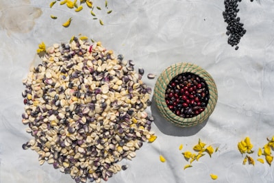 A collection of various seeds and grains spread on a light-colored surface, with a small woven basket filled with red and black seeds. Scattered around are yellow flower petals.