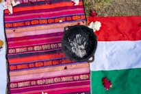 A colorful woven textile featuring intricate patterns in shades of orange, pink, and purple is spread out alongside a partially visible flag with red, white, and green sections. On top of the textile is a blackened pot containing ash or burnt material. Scattered flower petals are visible, adding a decorative element to the scene, which appears to be outdoors on grassy ground.