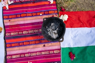 A colorful woven textile featuring intricate patterns in shades of orange, pink, and purple is spread out alongside a partially visible flag with red, white, and green sections. On top of the textile is a blackened pot containing ash or burnt material. Scattered flower petals are visible, adding a decorative element to the scene, which appears to be outdoors on grassy ground.