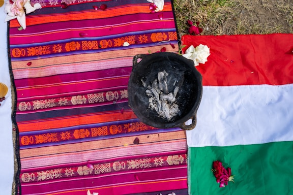 A colorful woven textile featuring intricate patterns in shades of orange, pink, and purple is spread out alongside a partially visible flag with red, white, and green sections. On top of the textile is a blackened pot containing ash or burnt material. Scattered flower petals are visible, adding a decorative element to the scene, which appears to be outdoors on grassy ground.