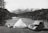 A serene outdoor scene features a vintage car parked next to a tent near a river with flowing water. In the background, snow-capped mountains rise above dense forests. A person is seated on the grass, relaxing and gazing at the natural landscape.
