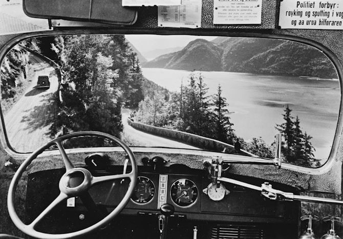 A cheerful tour bus driver smiling behind the wheel of his coach in a scenic European landscape.