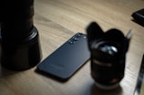 A smartphone with a sleek black finish and triple camera setup lies on a wooden surface, flanked by two camera lenses.