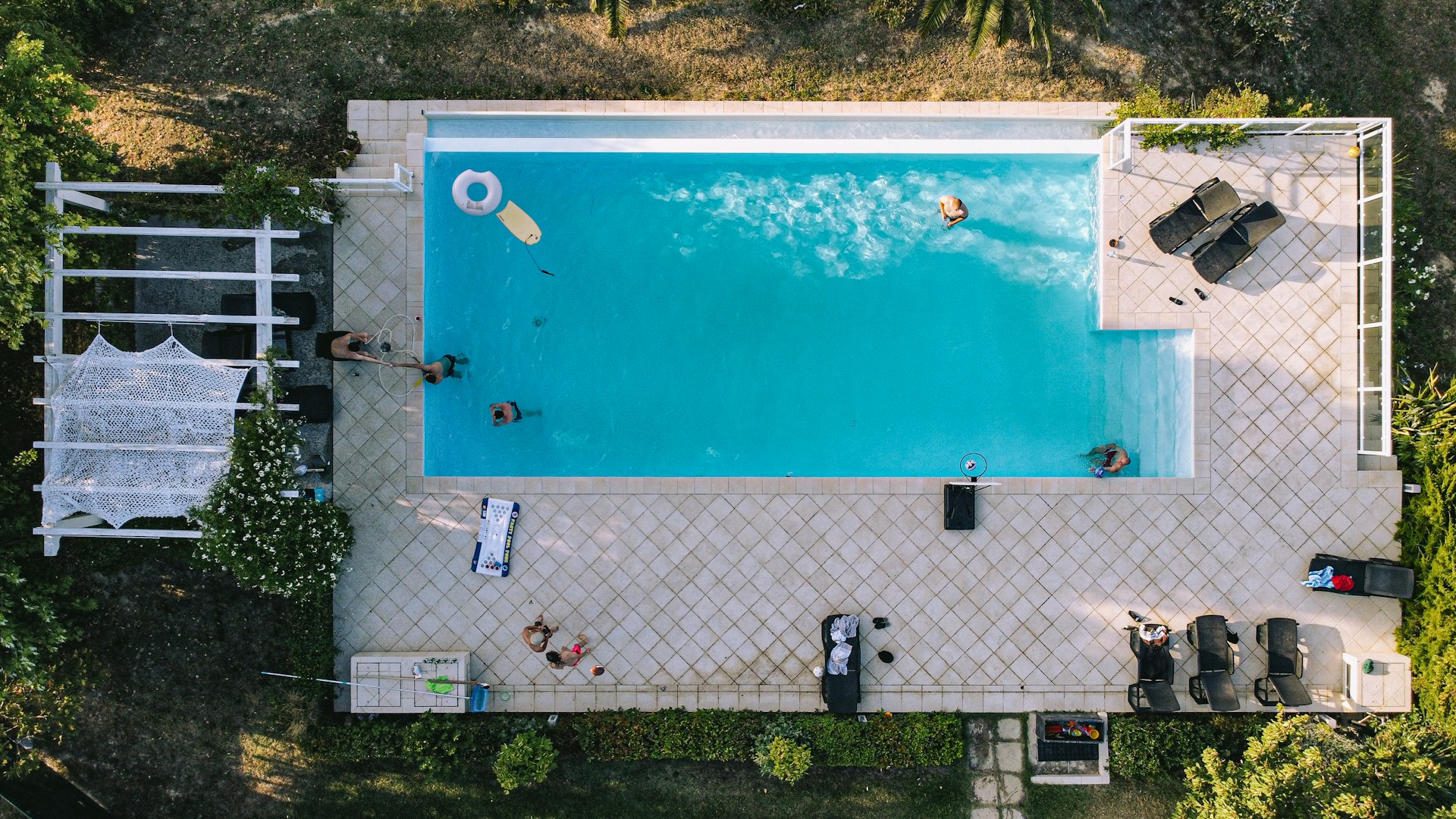 Aerial view of a swimming pool surrounded by a tiled deck. Several people are in the pool, enjoying leisure activities. There are sun loungers with towels and a shaded area with a trellis and greenery around the perimeter.