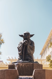 A statue of a small, wise creature with large ears and a robe is positioned atop a stone fountain. The surroundings include a clear blue sky and parts of buildings and trees in the background.