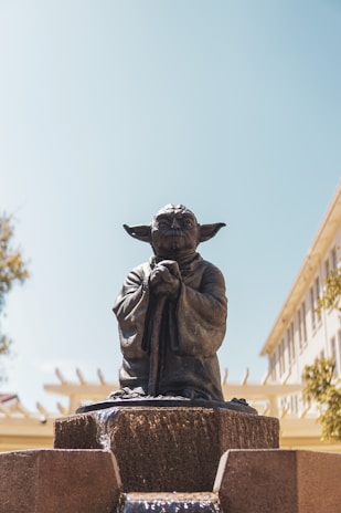 A statue of a small, wise creature with large ears and a robe is positioned atop a stone fountain. The surroundings include a clear blue sky and parts of buildings and trees in the background.