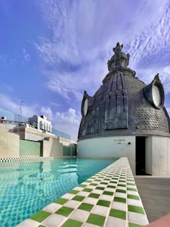 A rooftop setting featuring a swimming pool with green and white checkered tiles. Adjacent to the pool is a distinctive dome structure made of dark materials, adorned with ornate details and resembling an architectural feature. The sky is clear with scattered clouds, indicating a pleasant, sunny day.