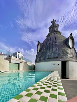 A rooftop setting featuring a swimming pool with green and white checkered tiles. Adjacent to the pool is a distinctive dome structure made of dark materials, adorned with ornate details and resembling an architectural feature. The sky is clear with scattered clouds, indicating a pleasant, sunny day.