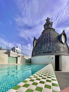 A rooftop setting featuring a swimming pool with green and white checkered tiles. Adjacent to the pool is a distinctive dome structure made of dark materials, adorned with ornate details and resembling an architectural feature. The sky is clear with scattered clouds, indicating a pleasant, sunny day.