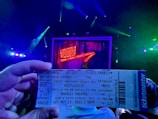 A hand holds a ticket for a theatrical show, with vibrant stage lights and a backdrop displaying a glowing sign. The atmosphere suggests excitement, with colorful lighting adding to the theatrical ambiance.