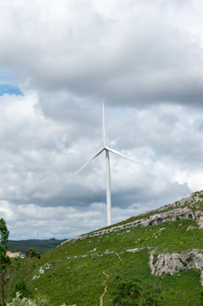 A large wind turbine stands on a grassy hill under a cloudy sky. The landscape features patches of greenery and rocky terrain, with a path leading to the turbine.