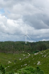 A wind turbine stands amidst a lush green landscape, surrounded by dense forests and hilly terrain. The sky is overcast with dense cloud cover, hinting at an impending storm or rain.