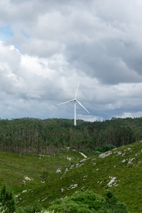 A wind turbine stands amidst a lush green landscape, surrounded by dense forests and hilly terrain. The sky is overcast with dense cloud cover, hinting at an impending storm or rain.