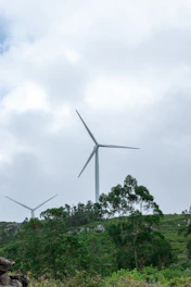Tall white wind turbines spinning gracefully on a green hillside against a bright, partly cloudy sky.